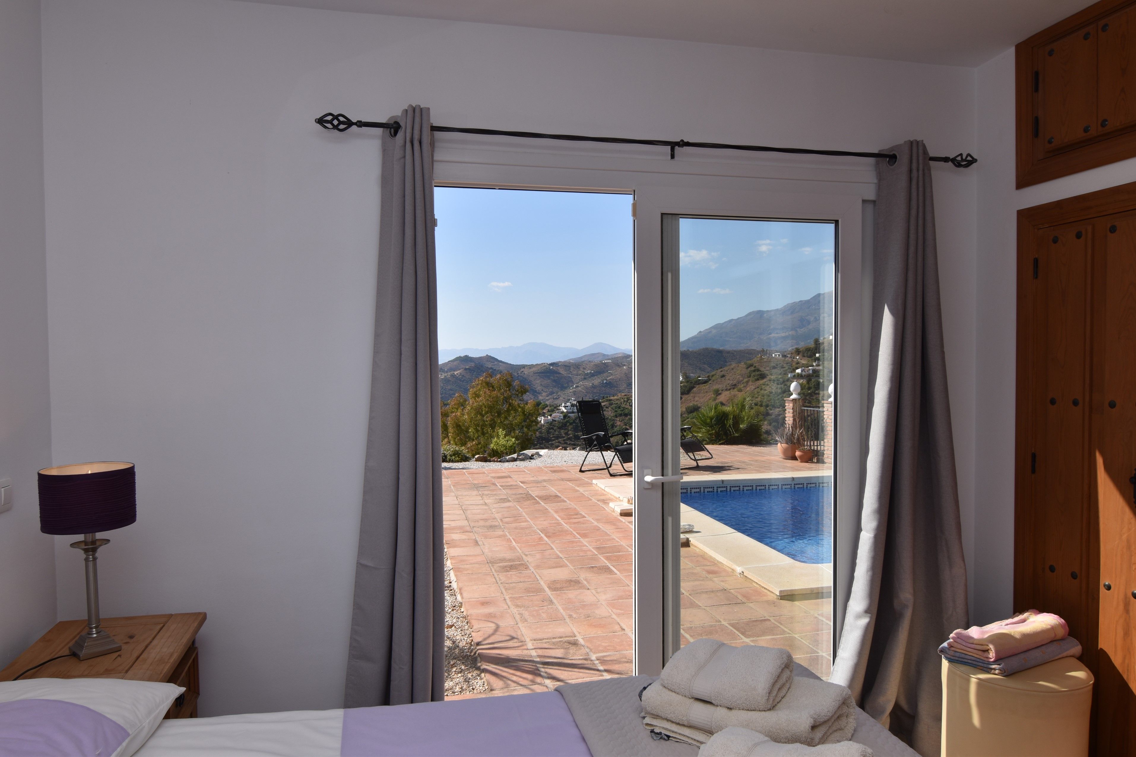 A bedroom with a bed, a nightstand, a lamp, a window with curtains, and a view of a pool and mountains.