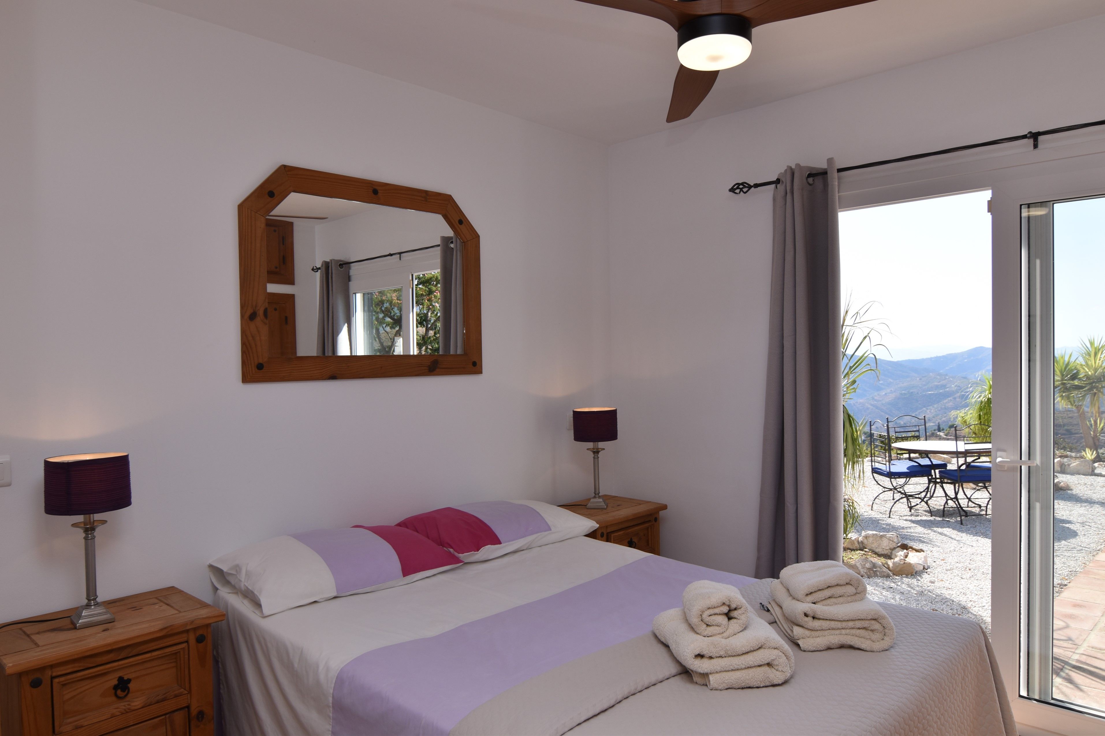 Une chambre à coucher avec un lit, des lampes de chevet, un ventilateur de plafond, un miroir et une vue sur l’extérieur.