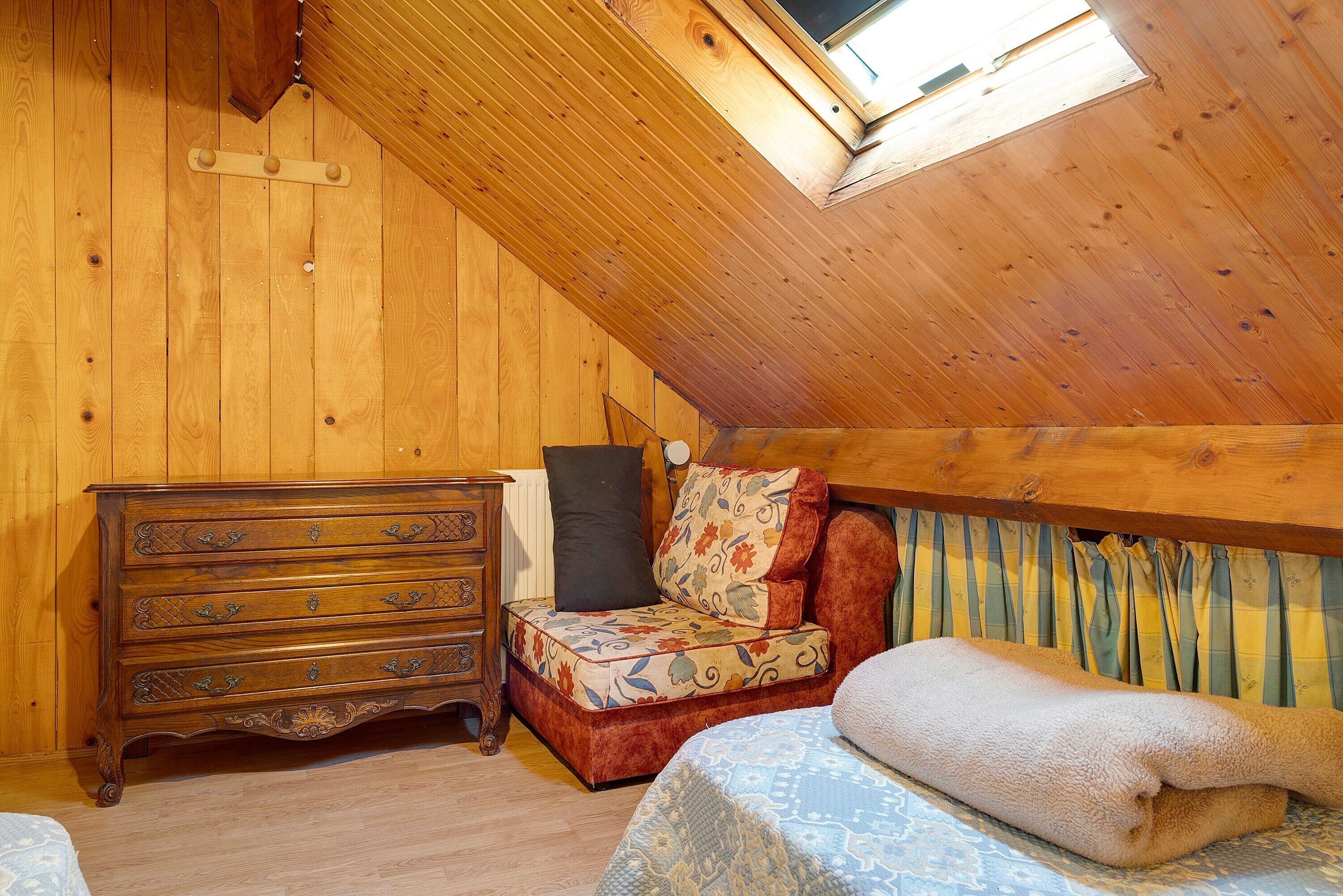 A wooden room with a sloped ceiling, a wooden dresser, a patterned chair, a bed with a blanket, and a window.