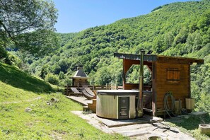 Una cabaña de madera con hidromasaje, mobiliario de exterior y vistas a una ladera boscosa.