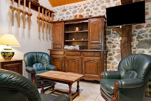 A room with a stone wall, wooden cabinets, leather chairs, a wooden table, and a television mounted on the wall. - Holiday Home 'Les Trois Maisons - Gite Vert' with Mountain View, Shared Pool and Wi-Fi (Osse-en-Aspe)