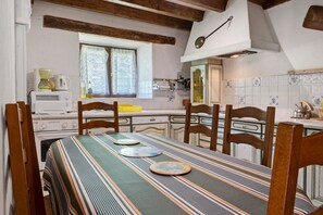 Ein Essbereich mit einem Tisch für vier Personen, Holzsesseln, einem Fenster mit Spitzenvorhängen, einem Herd und einer gefliesten Spritzschutzwand.