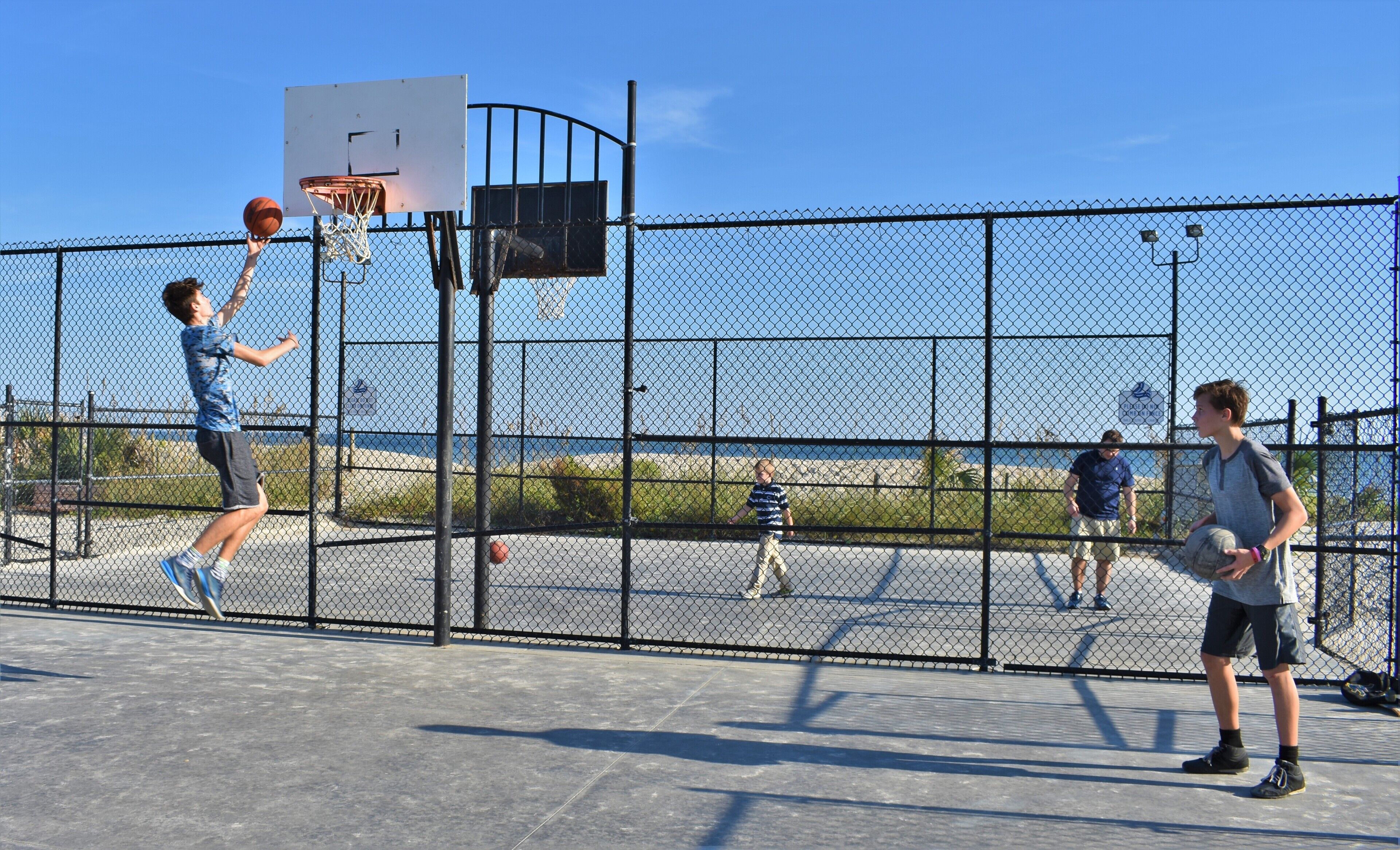 Een basketbalveld buiten, met een hek van gaas, een basketbalring en een helder uitzicht op de oceaan op de achtergrond.