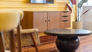 A round table with a dark base, a wicker chair, and wooden cabinets in the background. - Myo Design House: São Bento (Porto)