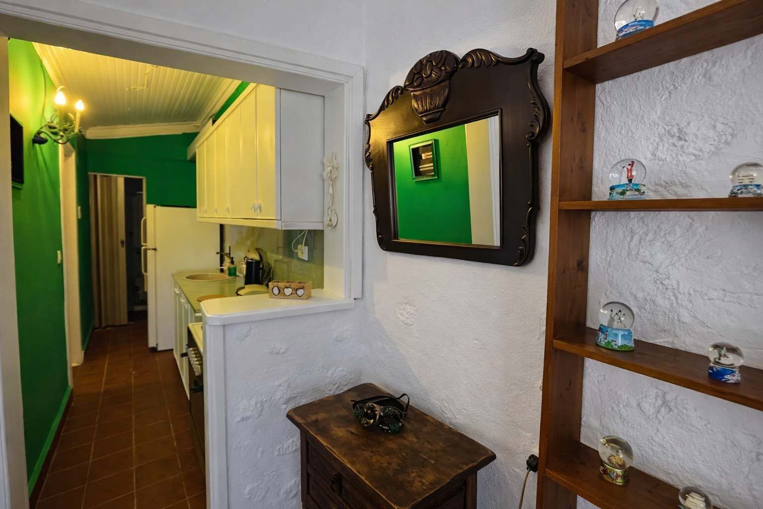 Um corredor estreito com parede verde, porta branca e um espelho com tela verde. Há uma pequena mesa de madeira com uma câmera e uma prateleira com objetos decorativos.