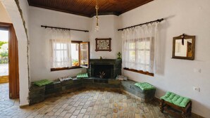 Une pièce avec une cheminée, des sièges confortables recouverts d’un tissu vert, un plafond en bois et des murs blancs.
