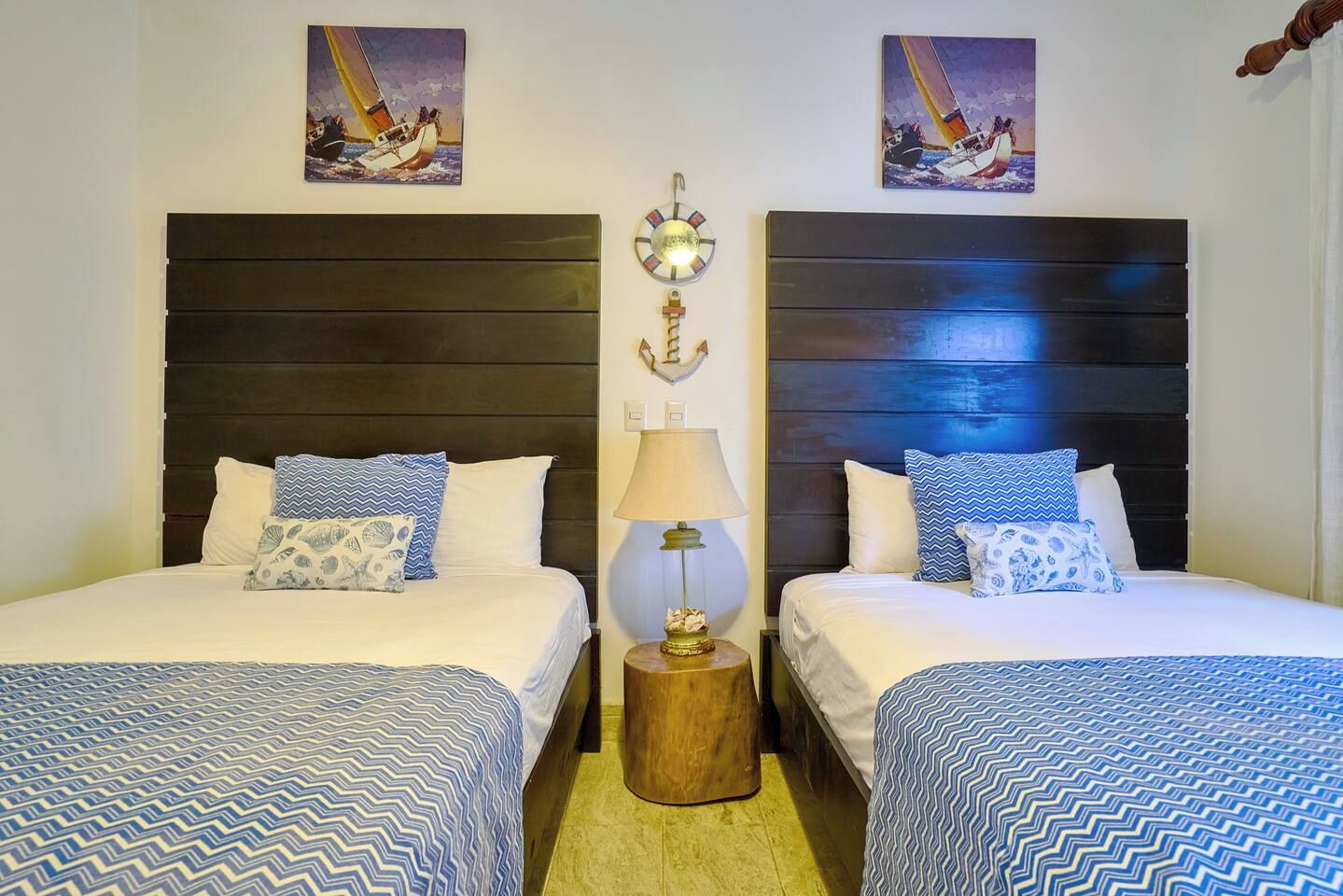 Quarto de hotel com duas camas, cada uma com colcha estampada, mesa de cabeceira com abajur e decoração de parede com tema náutico.
