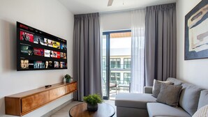 Ein modernes Wohnzimmer mit einem an der Wand befestigten Fernseher, der eine Netflix-Oberfläche anzeigt, einem Holztisch, einem runden Couchtisch mit einer Pflanze, einem grauen Sofa mit gemusterten Kissen und einem großen Fenster mit grauen Vorhängen.