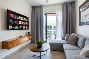 A modern living room with a mounted TV displaying a Netflix interface, a wooden console table, a round coffee table with a plant, a grey sofa with patterned cushions, and a large window with grey curtains. - Exclusive 1BR, pool, gym, and artificial beach (Punta Cana)