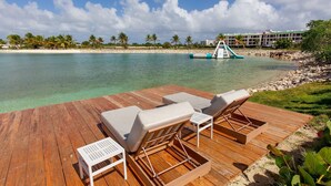Eine Holzterrasse mit Liegestühlen und einem kleinen Tisch, mit Blick auf ein klares türkisblaues Meer, einen von Palmen gesäumten Strand und eine Wasserrutsche im Hintergrund.