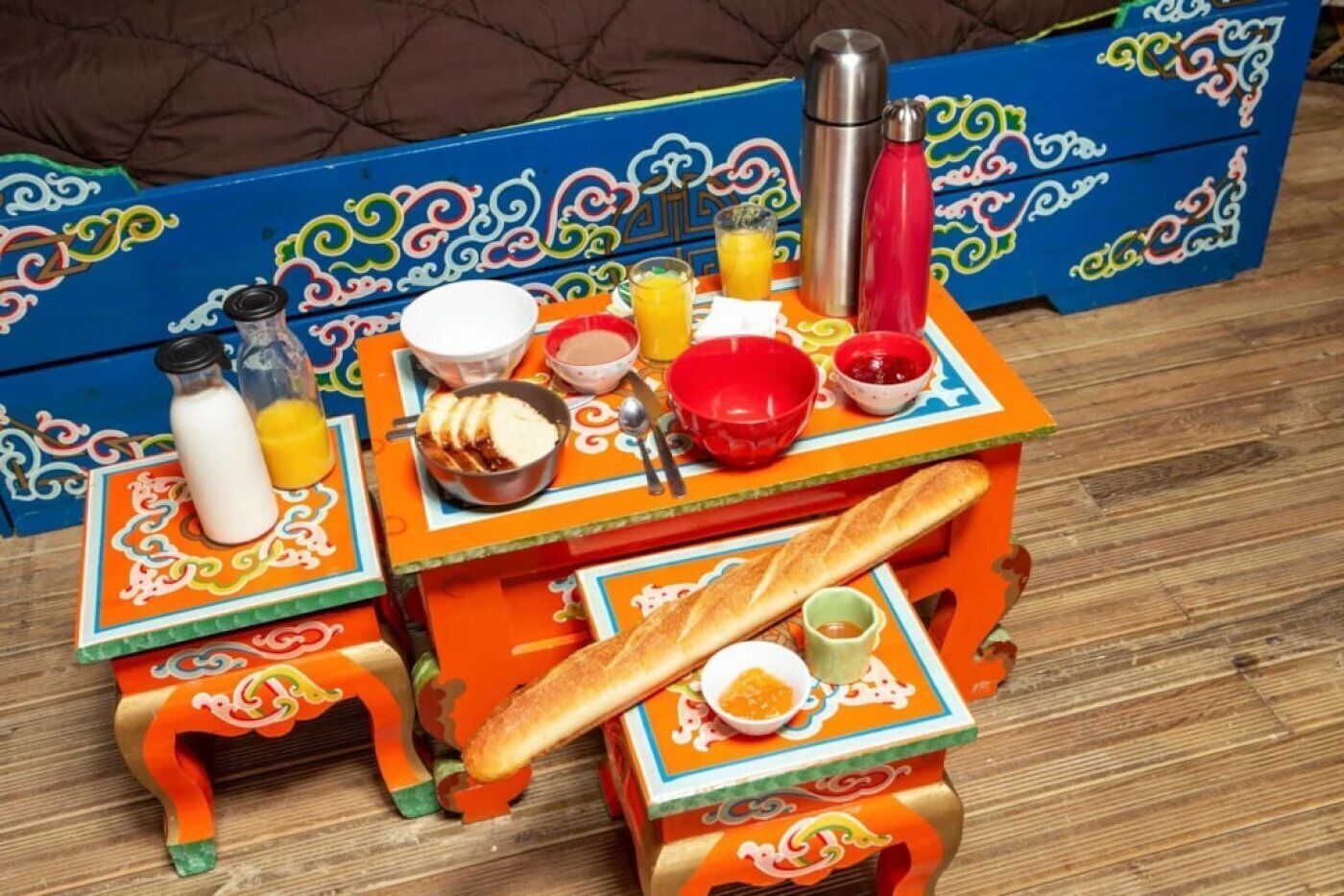 Una mesa colorida dispuesta con diversos alimentos para el desayuno, incluyendo pan, una botella de leche, un vaso de zumo de naranja y una termo.