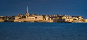 Un paisaje urbano costero con edificios históricos, una imponente aguja de iglesia y un muro fortificado a lo largo de la costa.