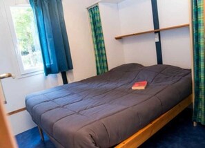 A single bed with a dark mattress and a red book on it, in a room with a window and curtains.