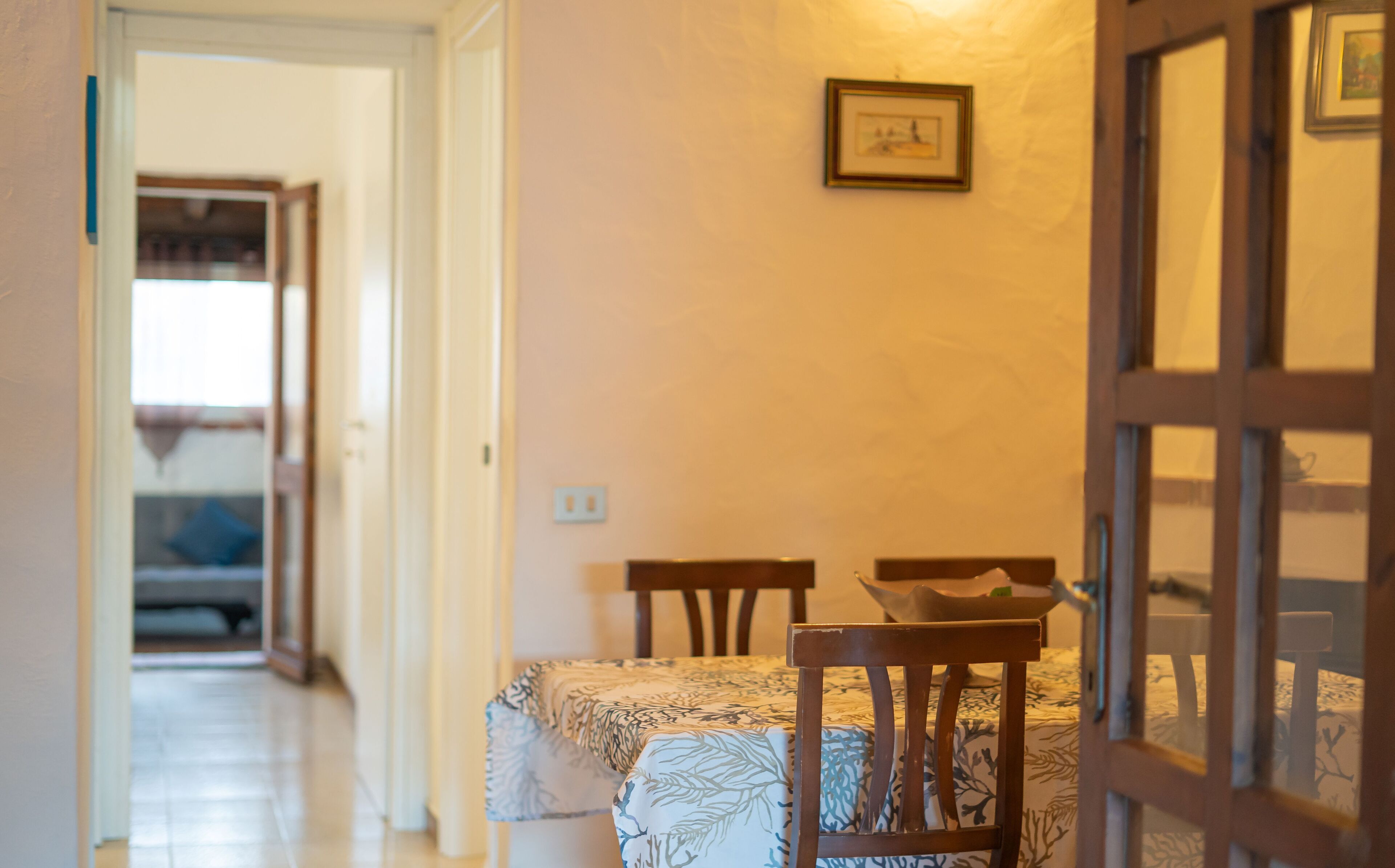 A hallway leading to a dining area with a table and chairs, a framed picture on the wall, and a door with glass panels.