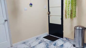 A hallway with a black door, a metal trash bin, a hanging plant, and a blue and white tiled floor.
