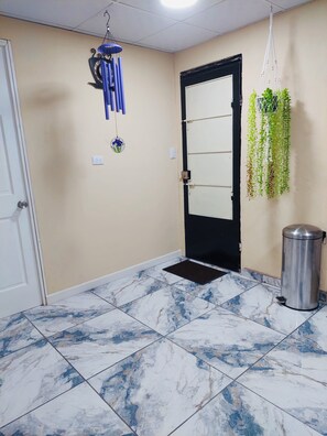 A hallway with a black door, a metal trash bin, a hanging plant, and a blue and white tiled floor.