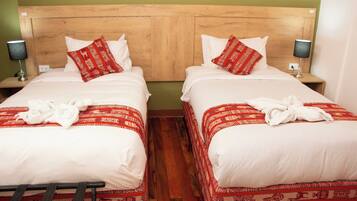 A hotel room with two beds, each with a wooden headboard, white bedding, and red patterned bed runners. There are two bedside tables with lamps, and a wooden floor.
