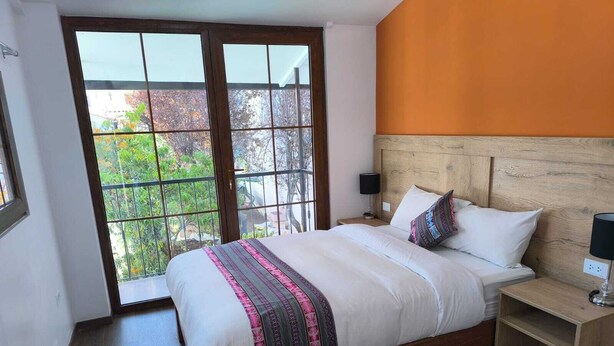 A bedroom with a large window, a wooden headboard, a bed with white bedding and a patterned throw, and a nightstand with a lamp. - san blas plaza hotel (Cusco)