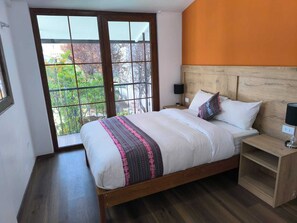 Un dormitorio con un ventanal grande, un cabecero de madera, una cama con ropa blanca y una manta estampada, y una mesita de noche con una lámpara.