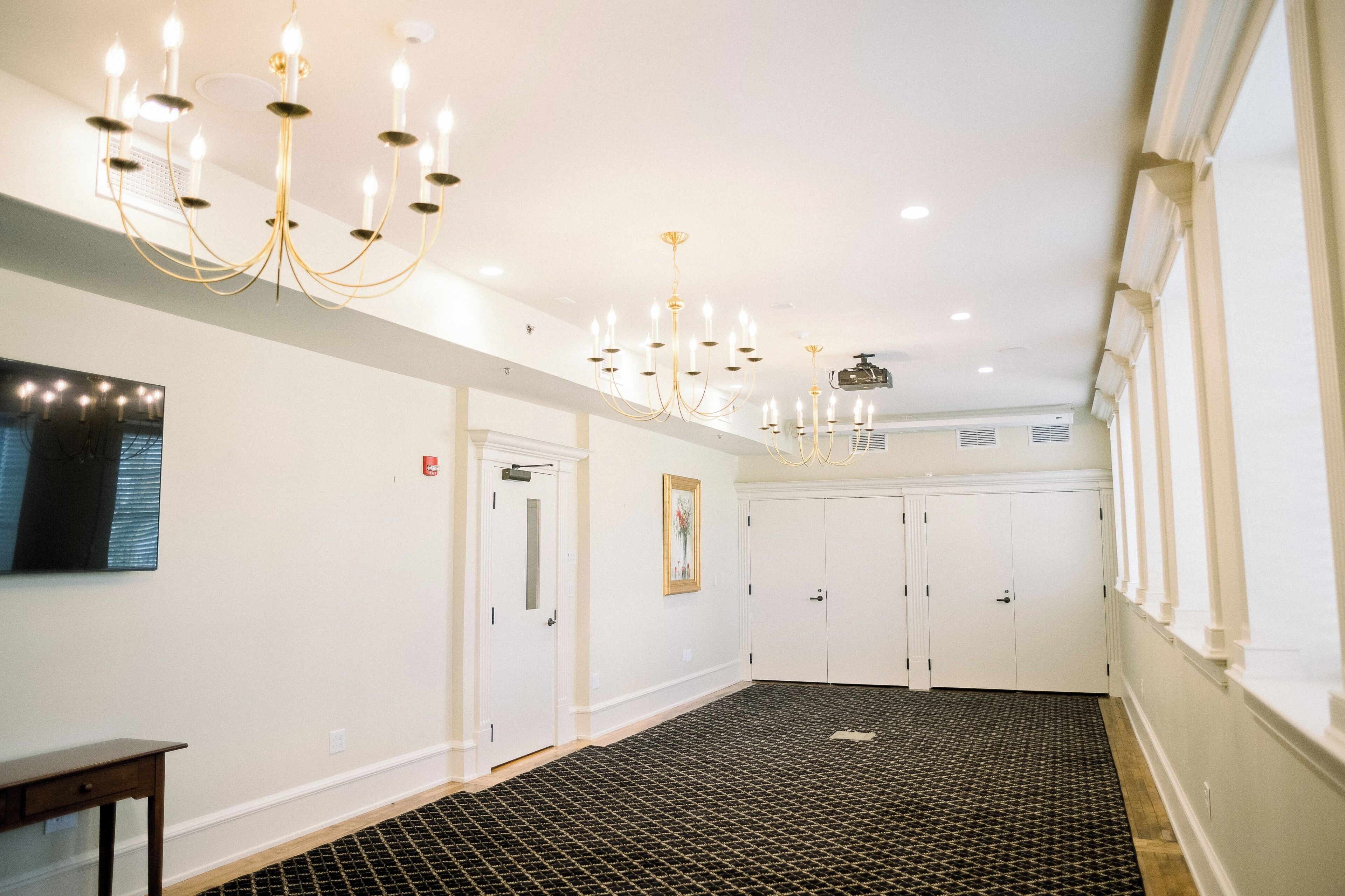 Un couloir d’hôtel avec des lustres, des murs blancs, une moquette à motifs et un banc en bois.