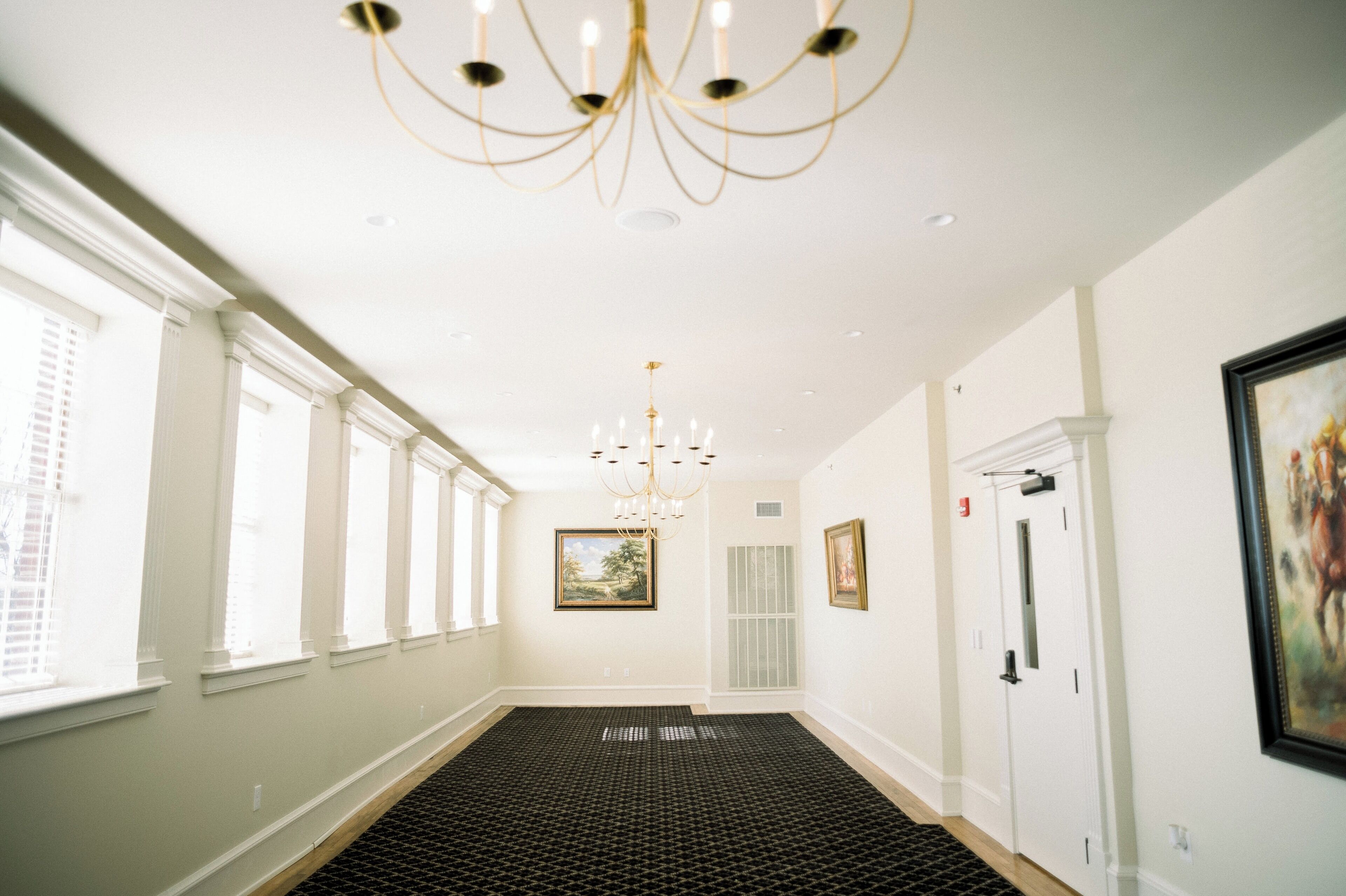 A hallway with a patterned carpet, a chandelier, framed artwork on the walls, and a door at the end.