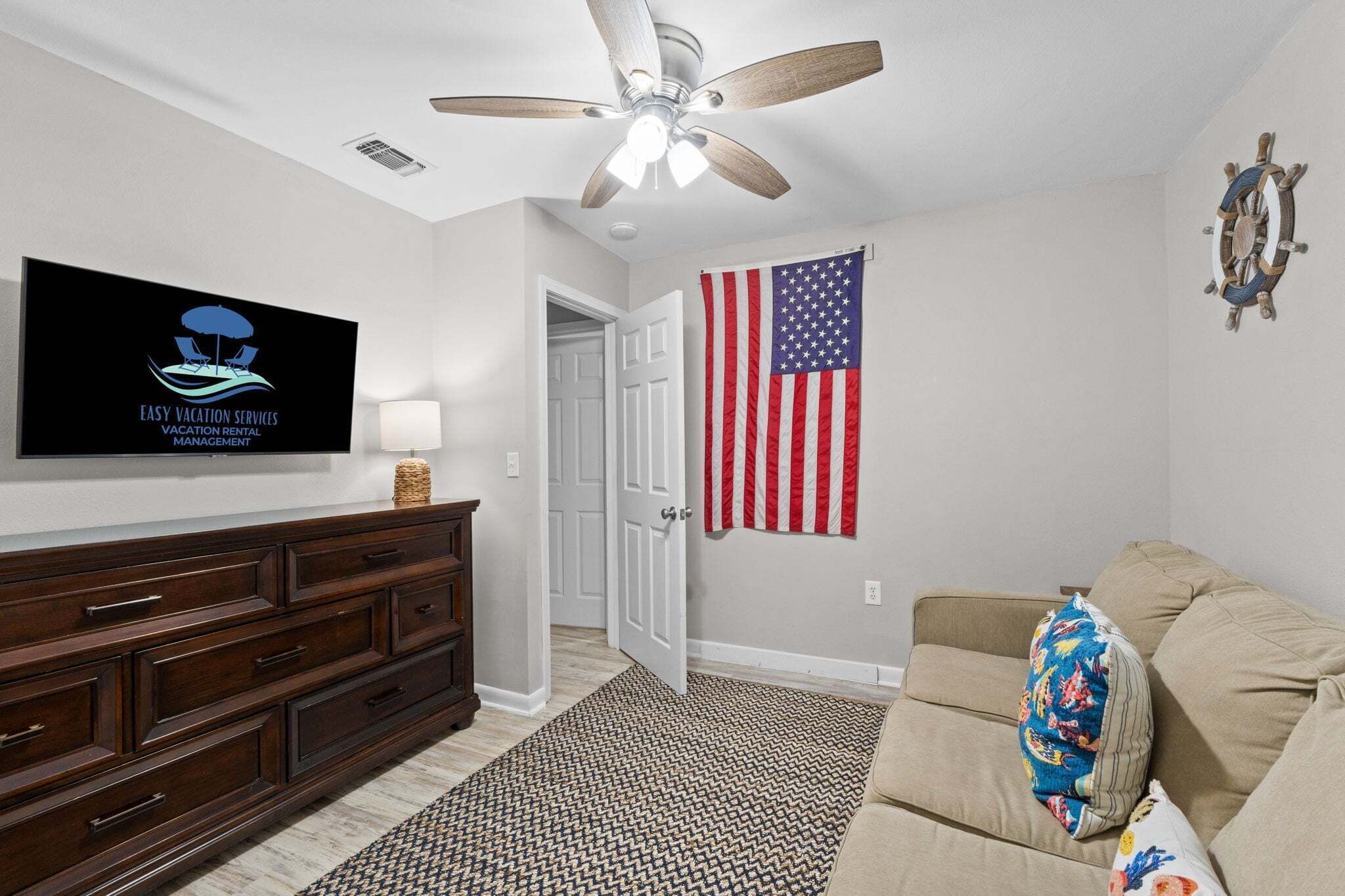 A room with a ceiling fan, a television displaying a logo, an American flag, a wooden ship decoration, a dark wooden dresser, and a beige sofa with a colorful pillow.