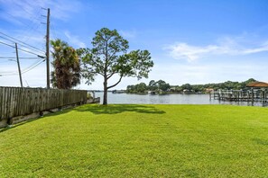 A grassy yard with a wooden fence, a large tree, and a body of water in the background. - Panhandle Paradise, Waterfront, 2 Dog Friendly in Fort Walton Beach. Free Activities Included! (Fort Walton Beach)