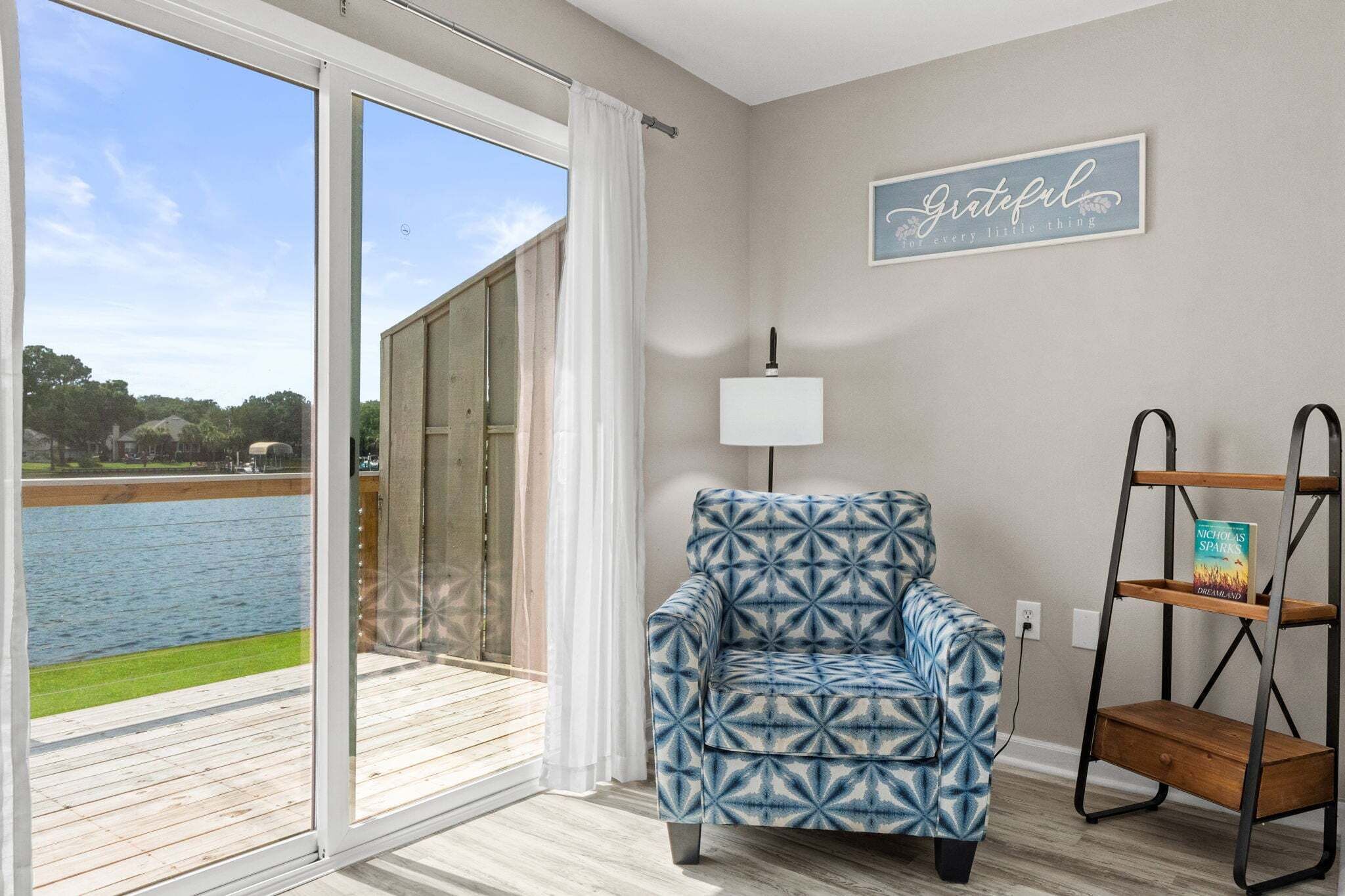 A room with a large sliding glass door leading to a deck, a patterned armchair, a wall-mounted lamp, and a shelf with a book.