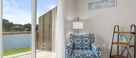 A room with a large sliding glass door leading to a deck, a patterned armchair, a wall-mounted lamp, and a shelf with a book.