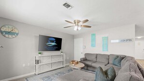 Een moderne woonkamer met een grijze bank, een flatscreen televisie, een plafondventilator en decoratieve wandkunst.