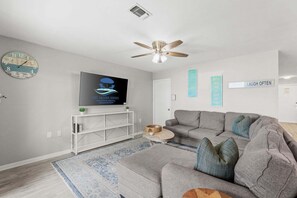 Ein modernes Wohnzimmer mit einem grauen Sofa, einem Flachbildfernseher, einem Deckenventilator und dekorativer Wandkunst.