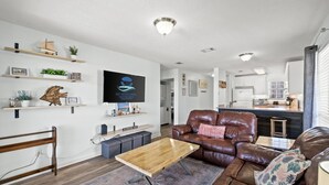 Un salon moderne avec un canapé en cuir marron, une table basse en bois et un téléviseur fixé au mur. On y trouve des étagères avec des plantes et des objets décoratifs, et on aperçoit un coin cuisine à l’arrière-plan.
