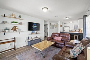 A modern living room with a brown leather sofa, wooden coffee table, and a TV mounted on the wall. There are shelves with plants and decorative items, and a kitchen area is visible in the background. - Walk to Beach! Free Activities! Coral Cove 301 (Fort Walton Beach)