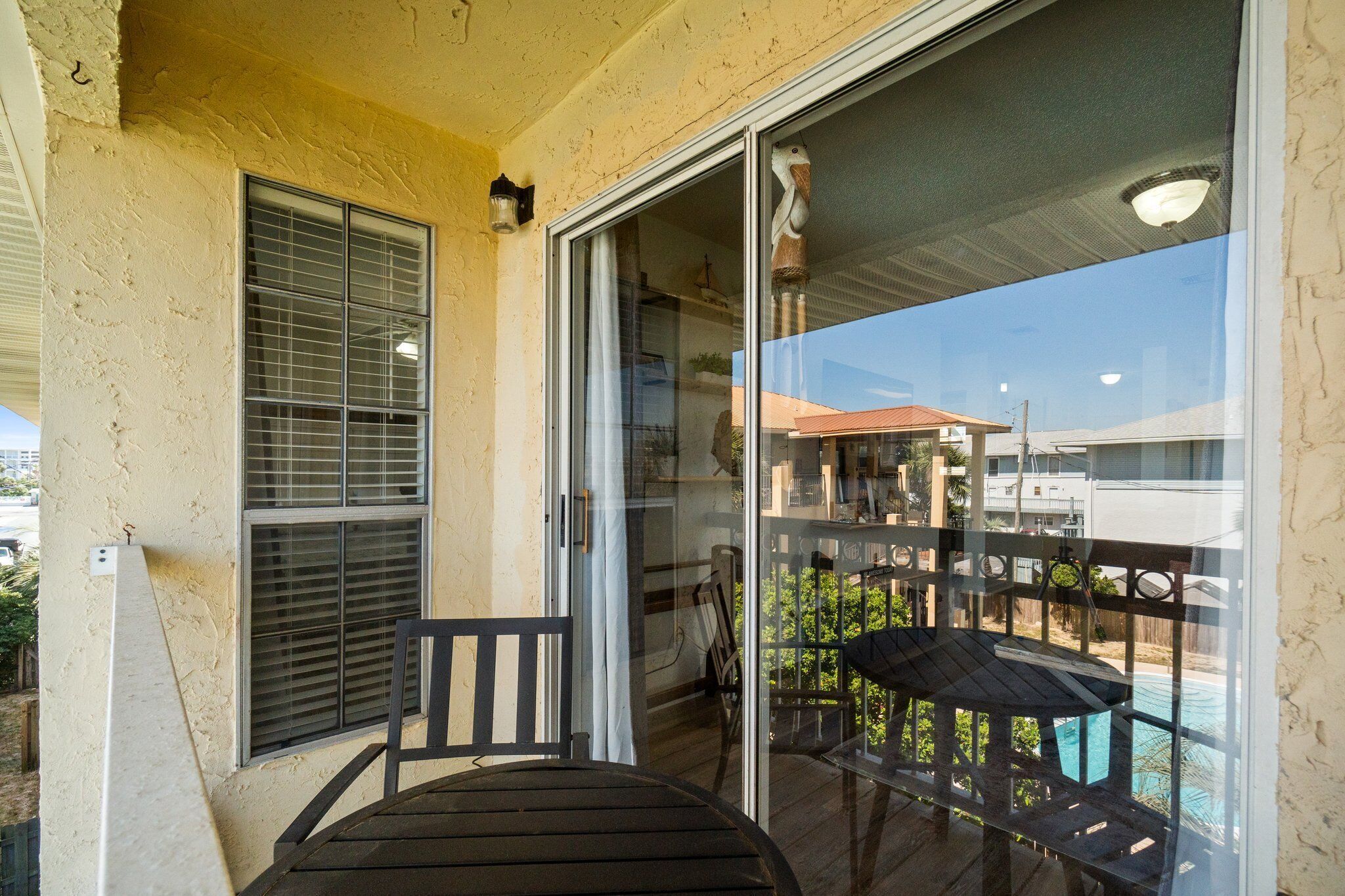 A balcony with a wooden table and chairs, a sliding glass door, and a view of a swimming pool and buildings in the background.