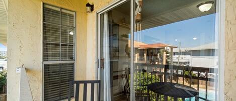 A balcony with a wooden table and chairs, a sliding glass door, and a view of a swimming pool and buildings in the background.