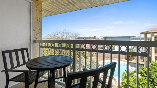 Un balcon avec une table ronde et deux chaises, donnant sur une piscine et les maisons voisines.