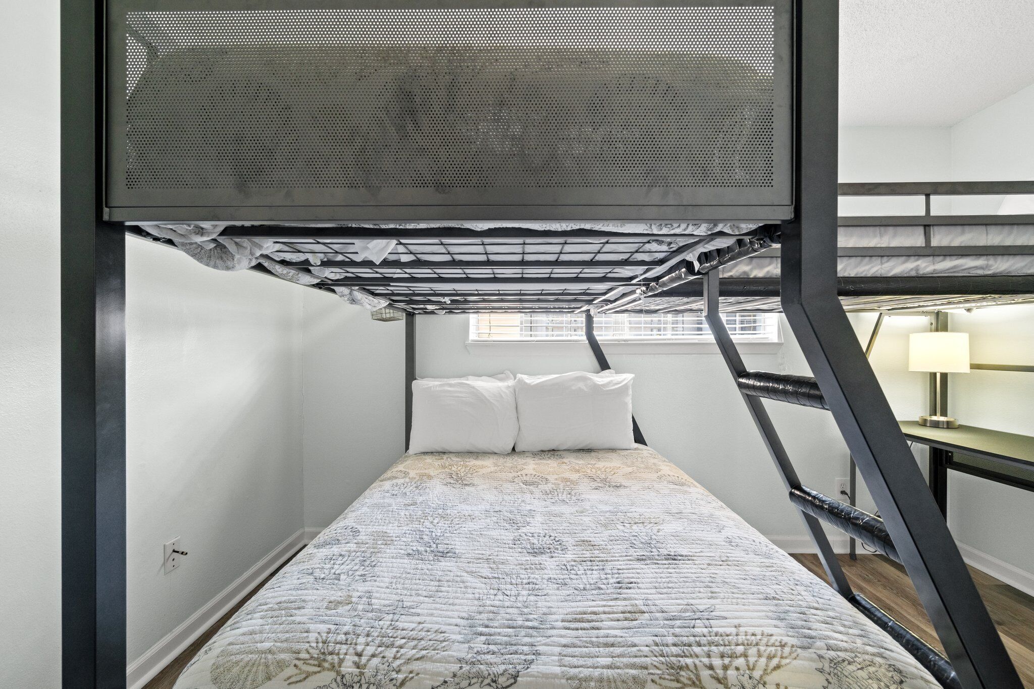 A compact room with a bunk bed, a small desk with a lamp, and a window providing natural light.