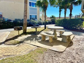 An outdoor patio area with a stone table and benches, surrounded by palm trees and a building with large windows. - Beachfront Condo with Pool, Hot Tub & Free Beach Service – Perfect for Families! (Fort Walton Beach)