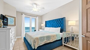 Quarto com uma cama grande, um ventilador de teto, mesa de cabeceira com luminária, cômoda e vista para o mar através de uma porta de correr.