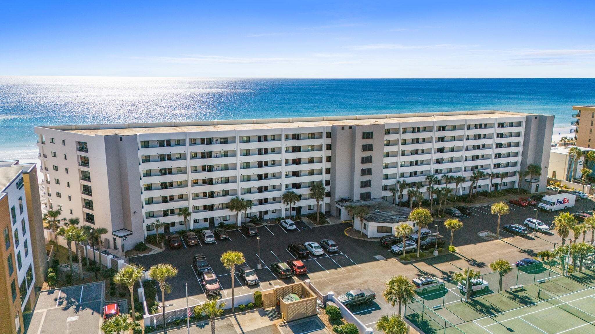 Een meerlaags hotel met een parkeerplaats, palmbomen en uitzicht op de oceaan.