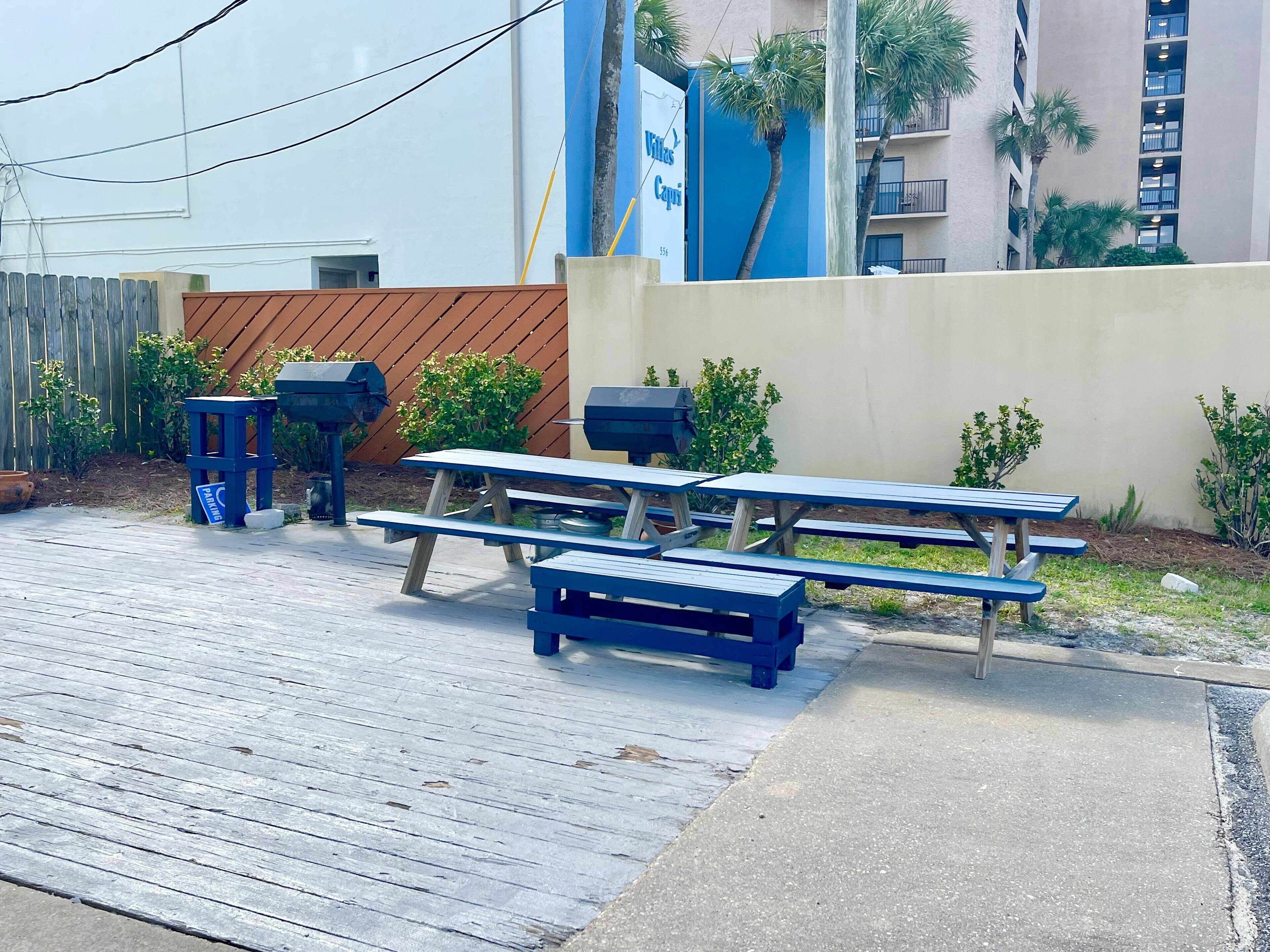 Des tables de pique-nique et des barbecues sont installés dans une cour agrémentée d’une clôture en bois et de quelques plantes.