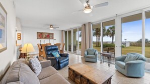 Ein großzügiges Wohnzimmer mit einer großen Glasschiebetür, die auf eine Terrasse führt. Es gibt einen Deckenventilator, einen Holztisch und mehrere Sofas und Sessel.
