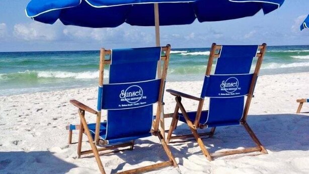 Kaksi rantatuolia, joiden yläpuolella on siniset aurinkovarjot. Aurinkovarjoissa on teksti »Sunset Beach Service Inc.». Tuolit ovat hiekkarannalla, ja taustalla näkyy valtameren aallot.