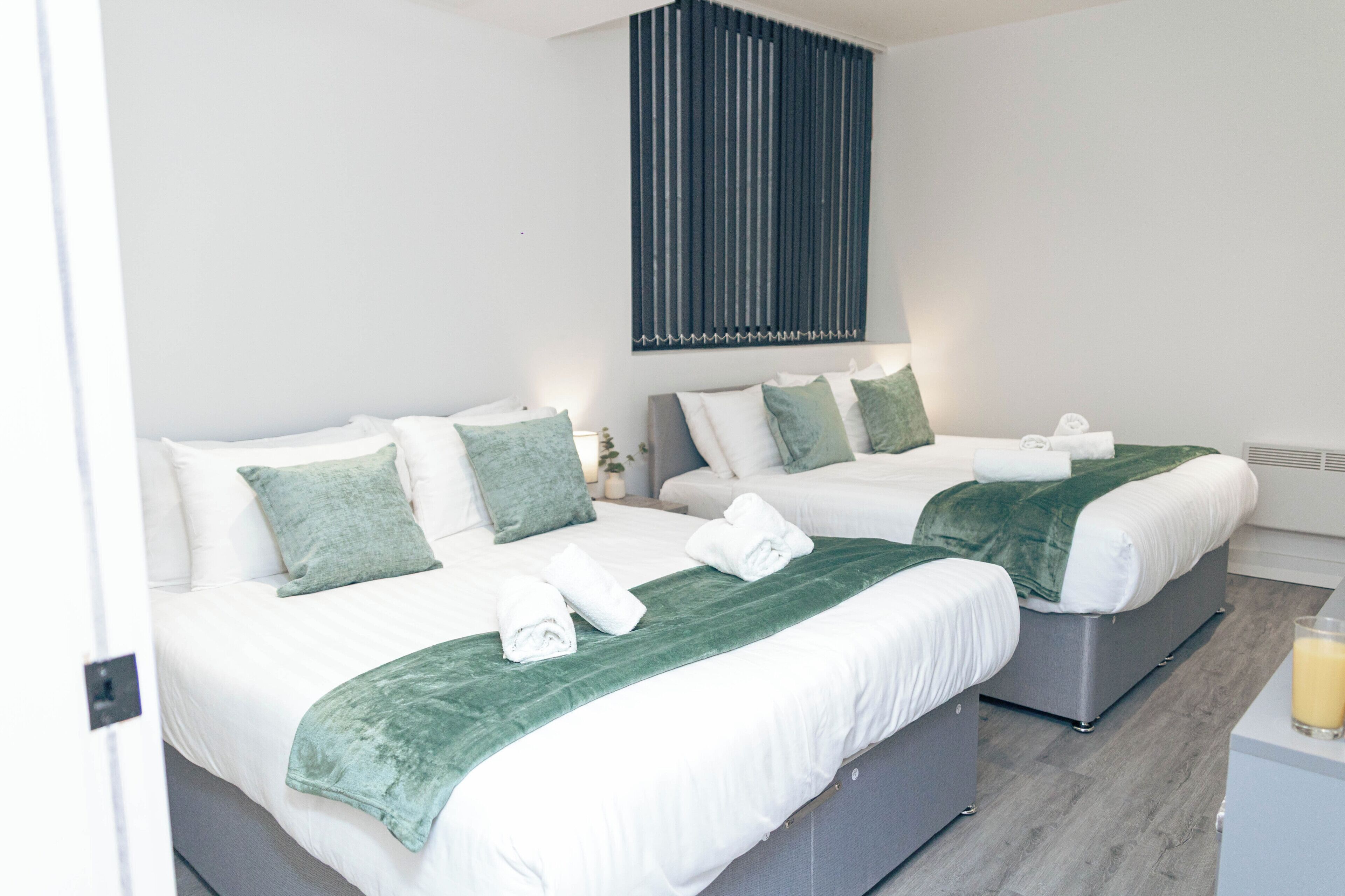 Habitación de hotel con dos camas, cada una con una manta verde y ropa de cama blanca. Encima de las camas, una lámpara de pared.