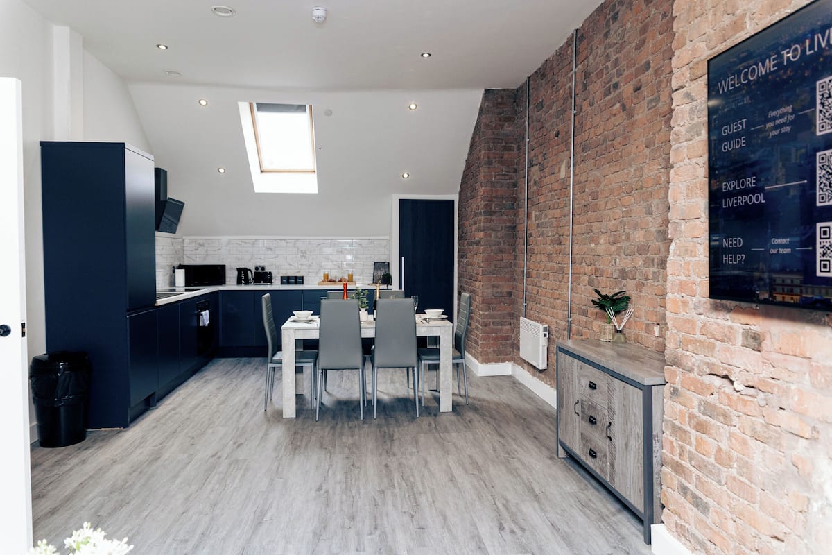A modern kitchen with dark cabinetry, a dining area with a table and chairs, and a brick wall with a welcome sign.