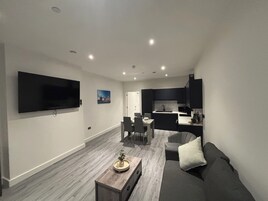 A modern living room with a flat-screen TV, a dining area, and a kitchen with dark cabinetry.