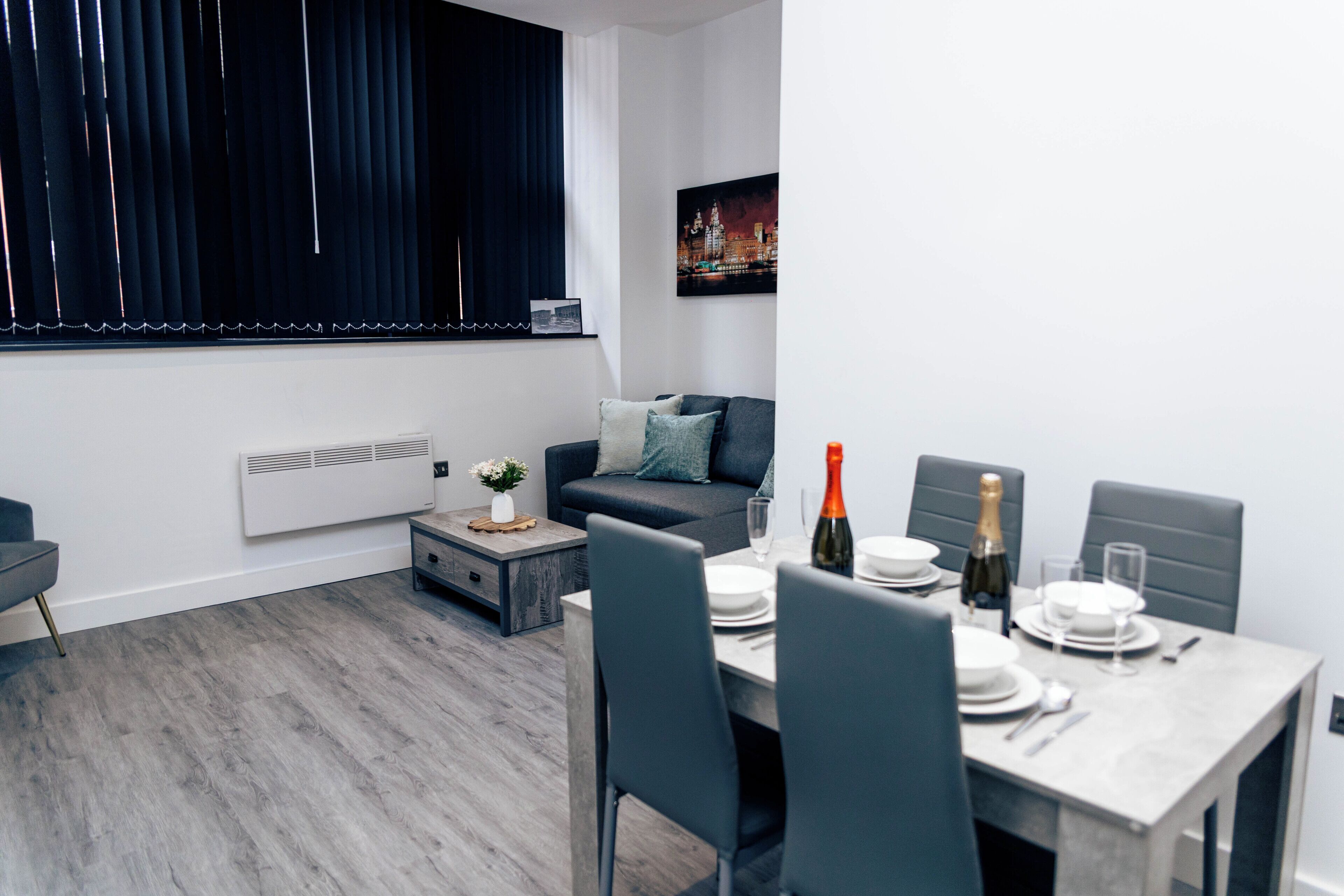 A modern living room with a sofa, a dining table set for two, and a window with blinds.