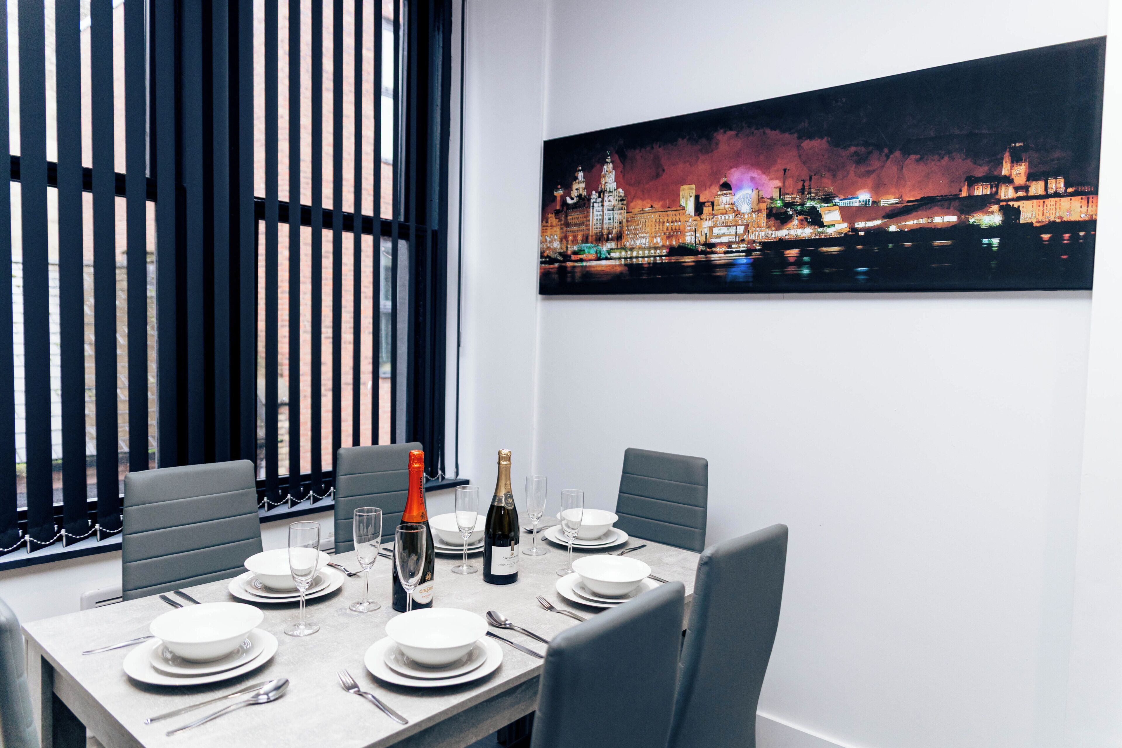 Mesa de jantar posta para seis pessoas, com um quadro de paisagem urbana na parede.