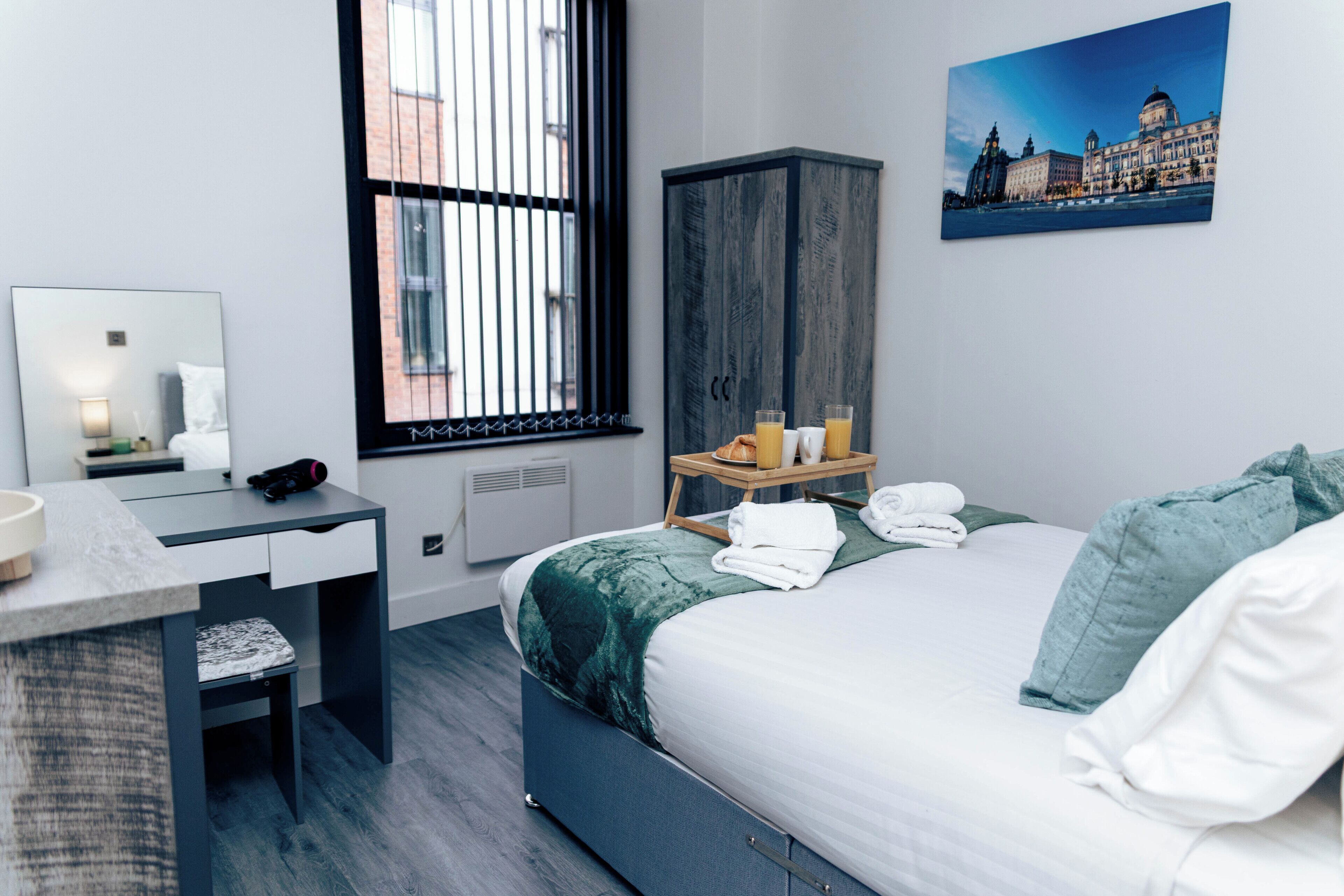 Une chambre d’hôtel moderne, équipée d’un lit, d’un bureau, d’une armoire, d’une fenêtre avec des stores et d’un tableau au mur.