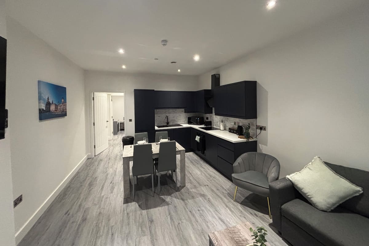 A modern kitchen with dark cabinetry, a dining area with a table and chairs, and a living area with a sofa and a plant.